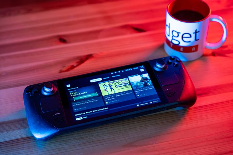 Ein Handheld-Spielgerät liegt auf einem Holztisch neben einer weißen Tasse mit dem Aufdruck 'Gadget Tester'. Das Display des Geräts zeigt eine Benutzeroberfläche mit Spielinformationen.