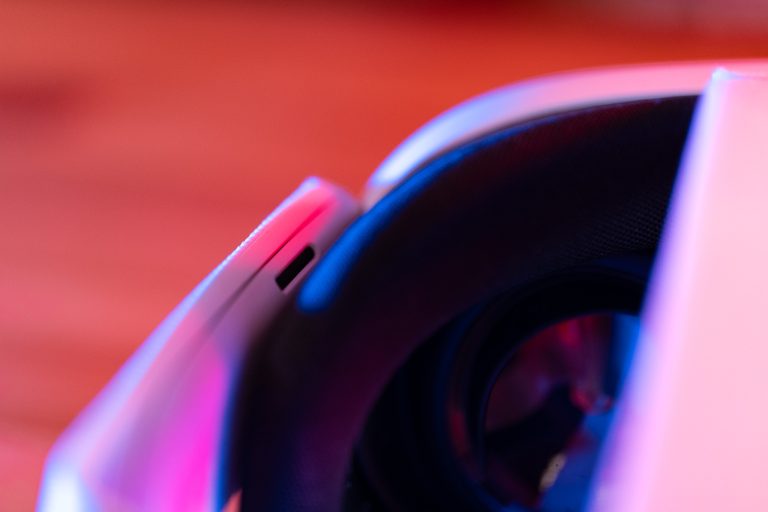 Nahaufnahme eines weißen technischen Geräts, möglicherweise eine VR-Brille oder ein Headset, mit einem Fokus auf das Polster und eine Linse. Der Hintergrund ist unscharf und in warmen Rot- und Pinktönen gehalten.