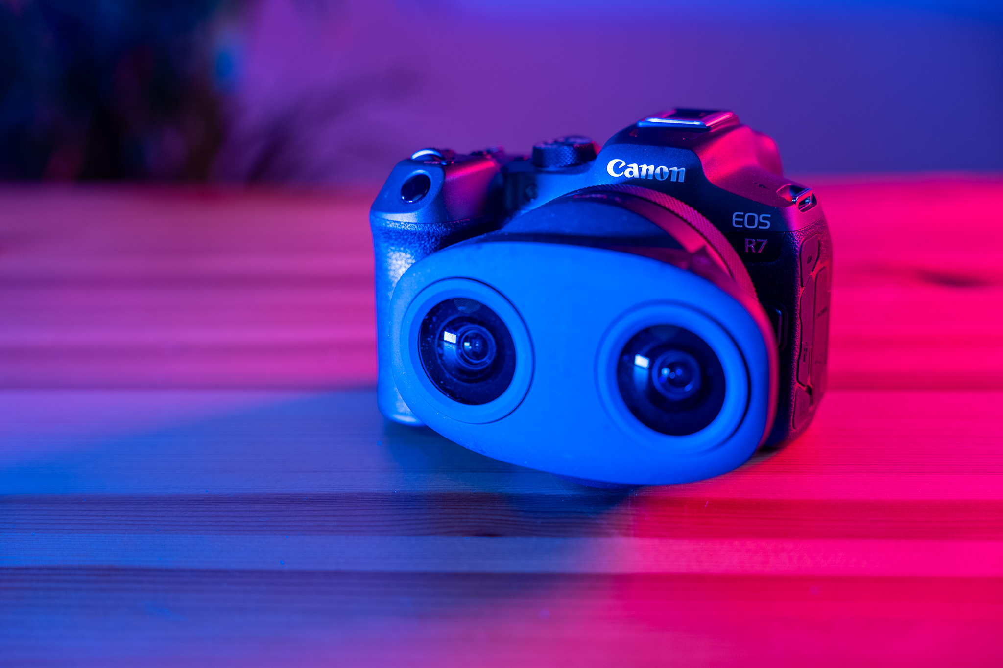 Eine Canon EOS R7 Kamera auf einem Holztisch, beleuchtet in Blau und Rosa. Zusätzlich ist ein blauer Adapter mit zwei Linsen angebracht.