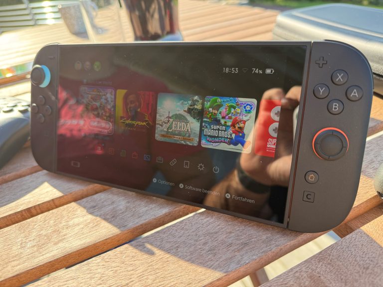 Eine Handheld-Spielkonsole auf einem Holztisch. Der Bildschirm zeigt eine Benutzeroberfläche mit mehreren Spielicons.