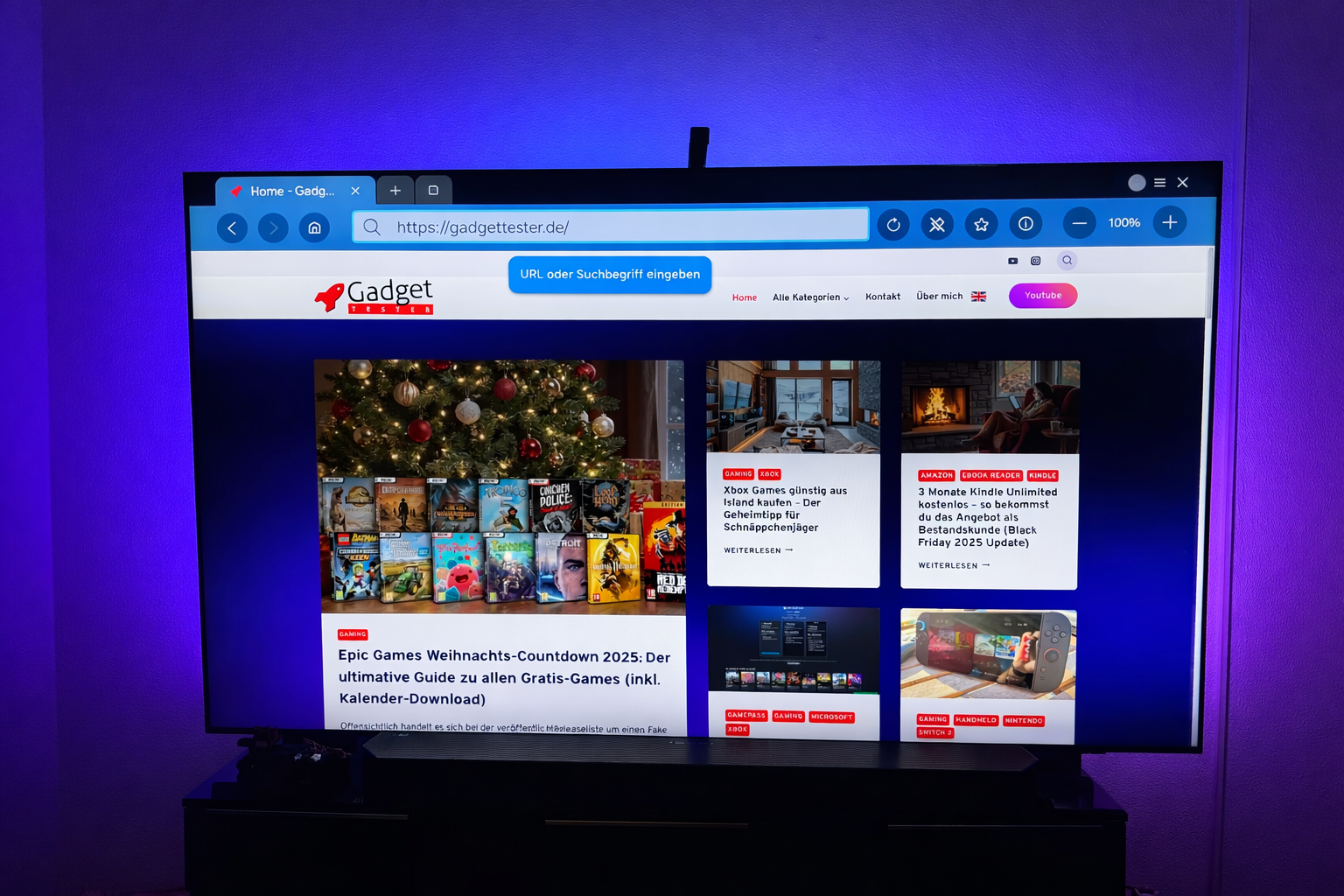 Ein Fernseher zeigt die Webseite von Gadget Tester mit Artikeln über Technik-Gadgets. Im Fokus steht ein Artikel über gratis Spiele von Epic Games, daneben verschiedene Technik-Themen.